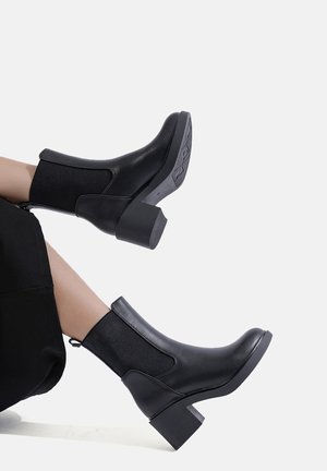 Stivaletti neri alla caviglia con finitura in pelle, pannelli laterali elasticizzati e tacco spesso. Texture liscia con una linguetta per facilitare l'indossamento.