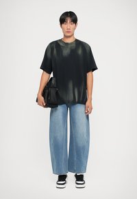 Must have must-valge T-särk, millel on plekiline muster, koos laiadest sinistest teksadega. Modelli käes on must kott ja jalas mustad tossud.