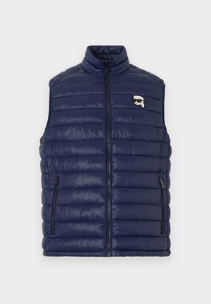 Tumesinine tikitud varrukateta vest esilukuga, kahe lukuga taskuga ja väikese logomärgiga, kus on päikeseprille kandva tegelase pilt.