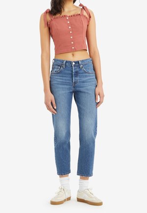 Femme portant un top court rose sans manches avec des boutons, un jean bleu taille haute, des chaussettes blanches et des baskets de couleur claire, debout devant un fond blanc.