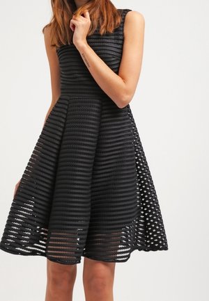 Mujer con vestido negro sin mangas hasta la rodilla con rayas horizontales texturizadas y falda acampanada, mano tocando la barbilla.