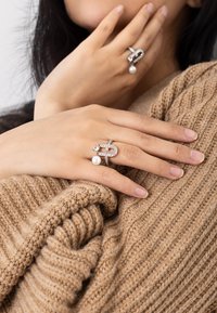 Anelli d'argento con pietre trasparenti e un accento di perla. Un anello presenta un design intrecciato; l'altro è una fascia con gemme che cadono.