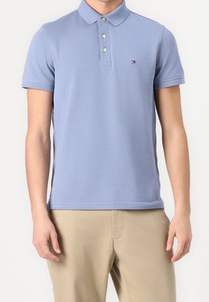 Man draagt een lichtblauw poloshirt met korte mouwen en een klein logo op de borst, gecombineerd met beige broek, staand tegen een effen achtergrond.