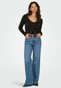 Young woman wearing a black fuzzy buttoned cardigan, blue high-waisted wide-leg jeans, brown belt, and black pointed shoes, standing with hands in pockets.