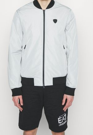 Mężczyzna noszący jasnoszarą kurtkę bomber z czarnymi mankietami i dołem, w połączeniu z czarnymi szortami z białym logo EA7 Emporio Armani.