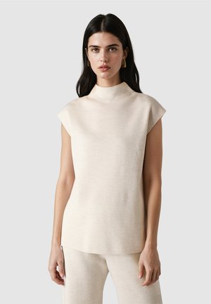 Mujer con cabello largo y oscuro lleva un top de punto sin mangas beige y pantalones a juego, de pie contra un fondo gris claro.