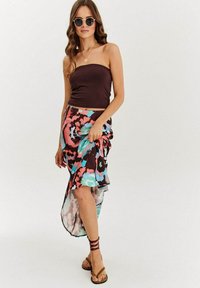 Hnedý top bez ramienok v kombinácii s voľnou sukňou s vysokým predkom a nízkym zadkom, ktorá má výrazný mnohofarebný abstraktný vzor. Hnedé sandále s obtočenými popruhmi.