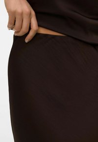 Ruskea kangas, jossa on sileä pinta. Käsi pitää reunasta lähellä materiaaliin tehtyä huomaamatonta viiltoa, esitellen yksinkertaisuutta suunnittelussa.