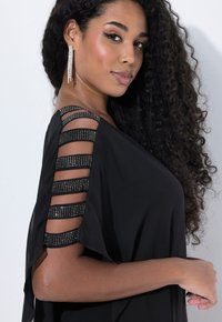 Vestido negro con un diseño asimétrico, tela translúcida y mangas rayadas adornadas con acentos metálicos. Cabello rizado y pendientes visibles.