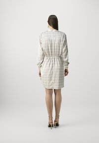 Femme debout de dos, portant une robe longue à manches longues beige à motifs avec une taille élastique et des sandales à talons hauts beiges sur un fond uni.