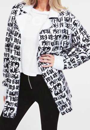 Zwart-wit cardi met een fuzzy textuur, voorzien van verschillende gedurfde gedrukte teksten en vormen. Draag met een witte top en zwarte broek.