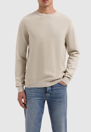 Beige sweatshirt met een textuur gebreid patroon, ronde halslijn en geribde manchetten, gecombineerd met lichtblauwe jeans.