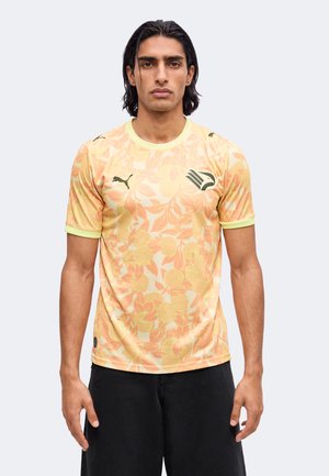 Jeune homme portant un t-shirt de sport jaune et orange à motif floral avec un short noir, debout, les bras détendus le long du corps.