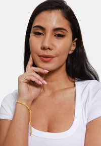 Bracciale in oro con slider regolabile, caratterizzato da una finitura liscia e una piccola perla di accento. Indossato sul polso sopra una maglia bianca.