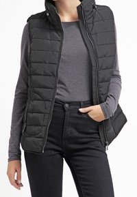 Černá péřová vesta s vysokým límcem, s horizontálním prošíváním, zapínáním na zip a bočními kapsami, nosí se přes šedou košili s dlouhým rukávem.