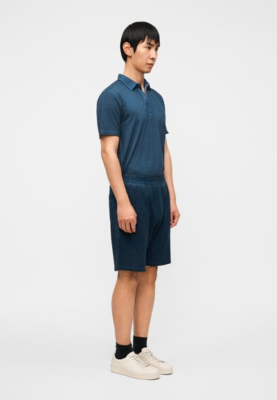 Μπλε σκούρο πόλο με γιακά και κουμπότρυπες, σε συνδυασμό με μπλε σκούρο σορτς. Απλός σχεδιασμός με άνετη εφαρμογή. Ανοιχτόχρωμα παπούτσια.