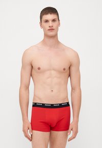 Röda boxerkalsonger med svart midjeband som har texten "HUGO". Tyget ser slätt och elastiskt ut, designat för en åtsittande passform.