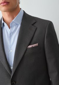 Dunkel strukturierter Blazer über einem hellblauen gestreiften Hemd mit weißen Streifen, das eine rosa Taschenquadrate aufweist. Der Blazer hat gekerbte Revers und eine Knopfverschluss.