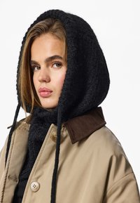 Bézs színű felsőruházat barna kordbársony gallérral, fekete kötött kapucnival párosítva. Texturált anyagokkal és laza szabású kialakítással rendelkezik.