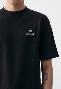 Čierne bavlnené tričko s okrúhlym výstrihom, s bielym vyšívaným logom a dizajnovými akcentmi na hrudi. Krátke rukávy.