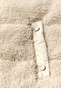 Tissu texturé de couleur crème avec une surface douce et pelucheuse ; présente une pièce de tissu lisse avec deux boutons-pression métalliques pour la fermeture.