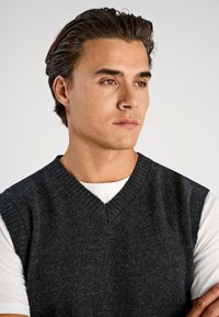 Grå ullväst med V-ringning, ribbad kant och ärmlös design bärs över en vit t-shirt. Slät textur med en klassisk passform.