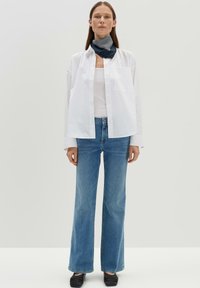 Witte blouse met knopen, blauwe denim wijde broek, zwarte instapschoenen en een marineblauwe gestreepte sjaal. Neutrale achtergrond benadrukt de outfit.