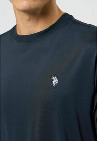 Tamsiai mėlynas trumpas rankovės marškinėliai, pagaminti iš lygaus audinio, su mažuoju baltu polo žaidėjo logotipu kairėje krūtinės pusėje.