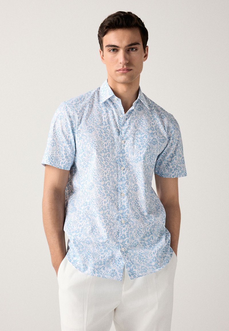 Rövid ujjú, világoskék anyagból készült, gombos ing virágmintával. Hegyes gallérral és enyhén laza szabással rendelkezik.