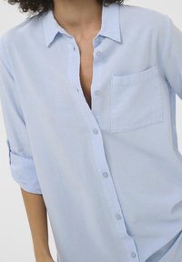 Femme portant une chemise boutonnée bleu clair aux manches retroussées et une poche sur le côté gauche de la poitrine, vue du cou jusqu'aux hanches.