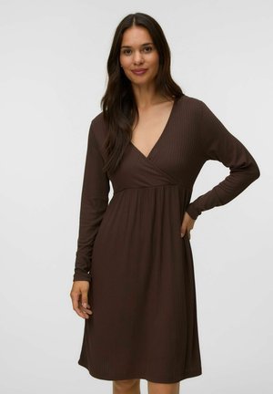 Femme aux longs cheveux foncés portant une robe marron à manches longues côtelée avec un col en V et une taille froncée, debout avec une main sur la hanche.