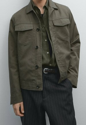 Homme portant une veste boutonnée vert olive sur une chemise assortie rentrée dans un pantalon foncé à fines rayures avec une ceinture tressée.