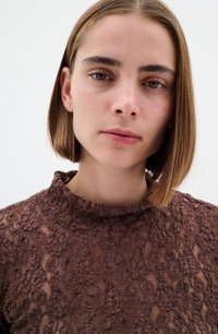 Top in pizzo marrone con collo alto e motivi floreali tessurizzati. Il tessuto appare trasparente con un design morbido e intricato.