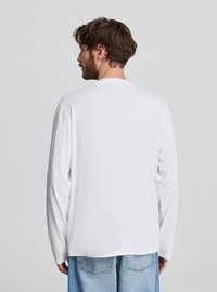 Biała koszula z długim rękawem, miękka bawełniana tkanina, okrągły dekolt i luźny krój. Noszona z jasnoniebieskimi dżinsami.