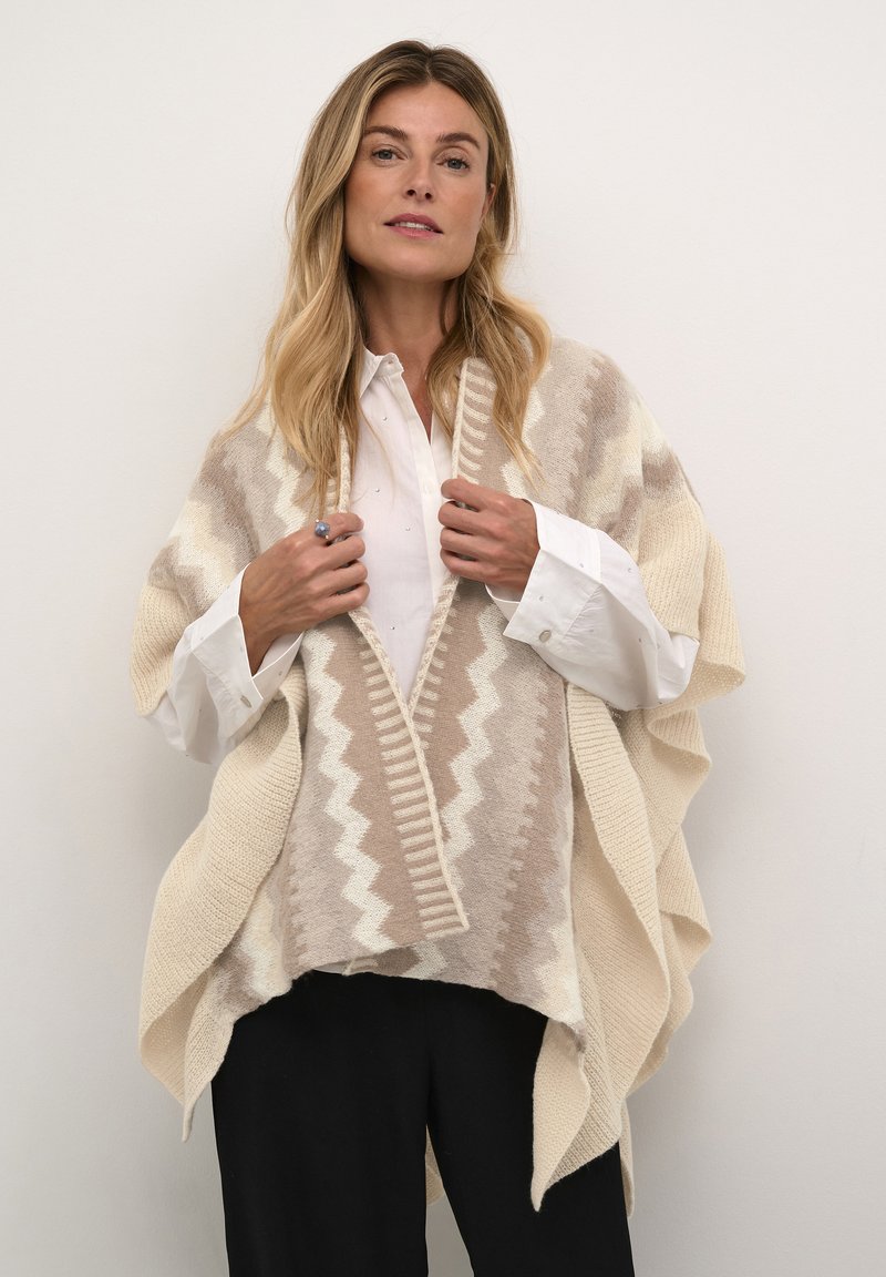 Beige og elfenben mønstret poncho med zigzag design, tekstureret strik, åben front og draperede ærmer, lagdelt over en hvid skjorte og sorte bukser.