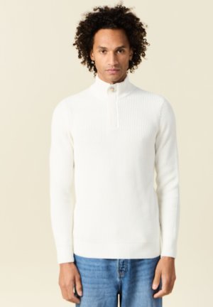 Weißer gerippter Pullover mit hohem Kragen und halblanger Reißverschluss. Aus weichem Stoff gefertigt, figurnaher Schnitt, kombiniert mit blauen Jeans.