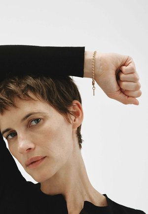 Persona con el cabello corto levantando el brazo, vistiendo una manga negra y una pulsera dorada de cuentas con un pequeño colgante en forma de llave sobre un fondo liso.