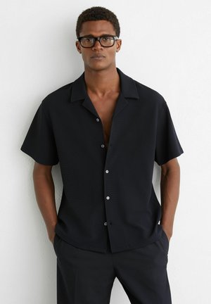 Mężczyzna w czarnej, krótkim rękawem koszuli zapinanej na guziki i okularach, stoi z rękami w kieszeniach przy białej ścianie.