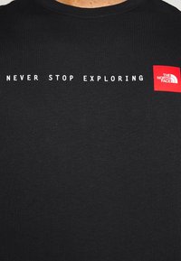 Camiseta negra con texto blanco "NUNCA DEJES DE EXPLORAR" y un logo cuadrado rojo de The North Face en la parte superior del pecho.