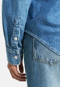 Jeansskjorte med lys blå vask, med knappelukking på ermene og lommedetaljer på baksiden. Glatt tekstur og uformelt design.