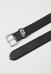 Ceinture en cuir noir avec une boucle rectangulaire en argent, présentant un logo circulaire. Texture lisse avec cinq trous d'ajustement.