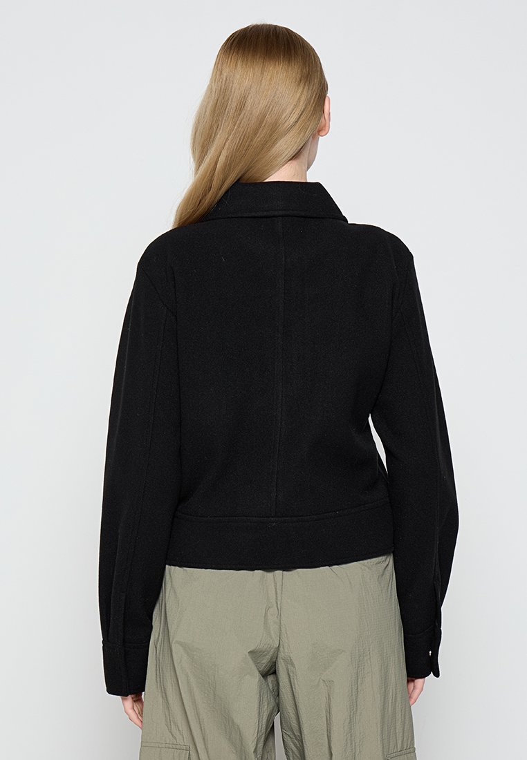 Veste noire en mélange de laine avec un col structuré, une taille ajustée et des manches longues. Présente une texture lisse et des détails de design ajustés.