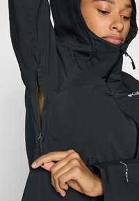 Black waterproof jacket with a hood, featuring zippered side pockets and textured fabric. Includes adjustable features and brand logo on sleeve.