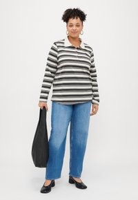 Melnbaltās svītrainās garumsvītru polo krekls ar gaišu apkakli un trim pogām, kopā ar gaiši ziliem taisnajiem džinsiem un melnajām kurpēm.