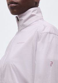 Κοντινό πλάνο ενός ατόμου που φορά ένα ανοιχτόροζ μπουφάν με ψηλό γιακά, φερμουάρ και διακριτικό λογότυπο στο στήθος και το μανίκι.