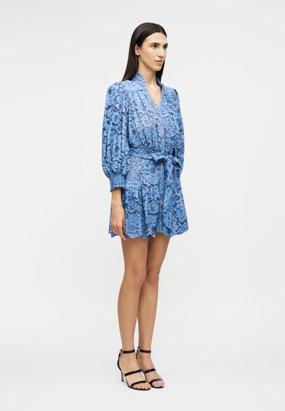 Woman standing wearing a blue floral lace mini dress with long puff sleeves, a waist tie, and black high-heeled sandals.