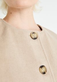 Cappotto in misto lana beige con scollo rotondo, caratterizzato da un evidente dettaglio di bottone in un colore a contrasto e texture liscia.