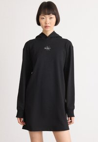 Čierne šaty s kapucňou vyrobené z hladkej tkaniny, s dlhými rukávmi a malým logom Calvin Klein na hrudi. Jednoduchý a minimalistický dizajn.