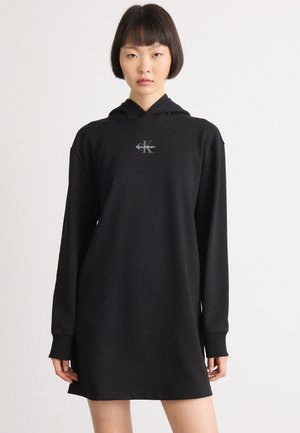 Crna haljina s kapuljačom izrađena od glatkog materijala, s dugim rukavima i malim Calvin Klein logotipom na prsima. Jednostavan i minimalistički dizajn.