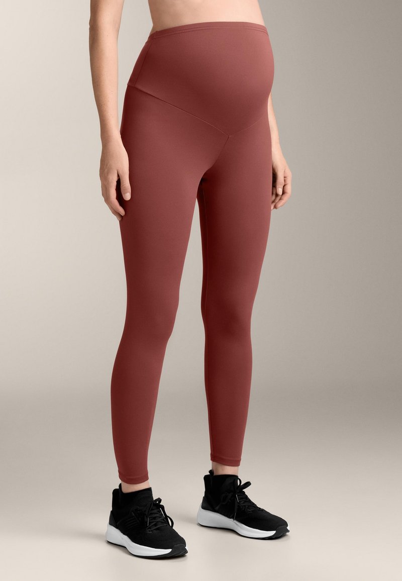 Personne enceinte portant un legging de maternité couleur rouille et des chaussures de sport noires sur un fond neutre.